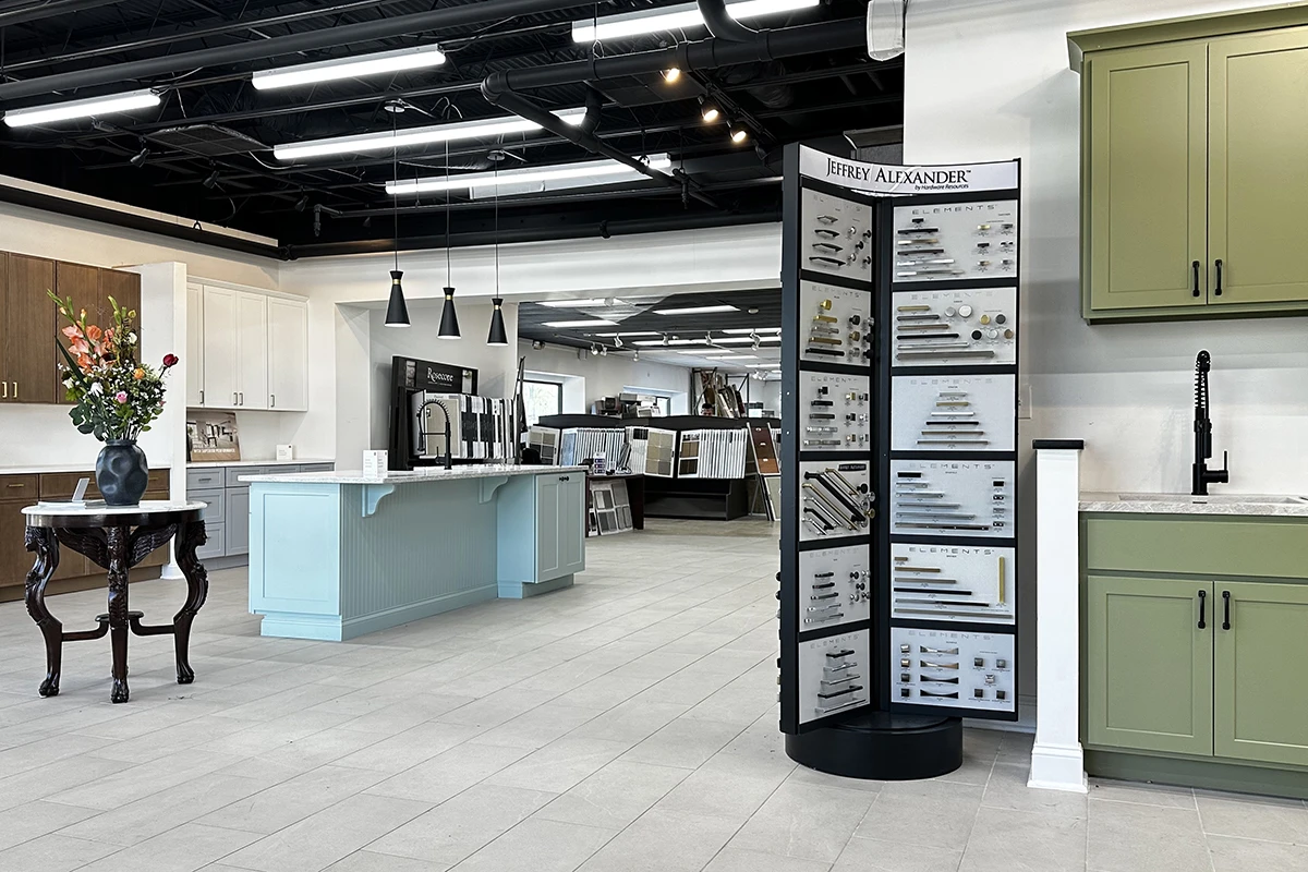 Kitchen cabinet showroom featuring white, blue, and sage green display cabinets, countertop samples, sinks, and a central hardware selection tower.